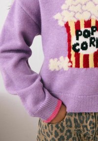 Lila gestrickter Pullover mit einem Popcorn-Graphic, mit gerippten Bündchen in Pink. Kombiniert mit Leopardenmuster-Hosen. Weicher Stoff, lässige Passform.