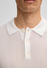 Polo bianco con tessuto strutturato, dotato di colletto classico e tre bottoni perlati. Piccolo foro evidente vicino al colletto.
