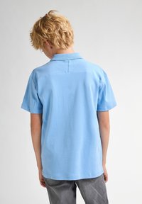 Ljusblå bomullspolo med krage, korta ärmar och rak nederkant. Har en slät yta och enkel design på baksidan.