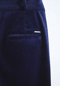 Pantalon en velours côtelé bleu marine montrant une poche arrière avec une petite étiquette de marque en métal rectangulaire sur le coin supérieur droit.