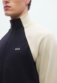 Vetoketjullinen takki, jossa on tummansininen vartalo ja vaaleanväriset hihat, valmistettu teksturoidusta kankaasta ja jossa on pieni kirjailtu "BOSS"-logo rinnassa.