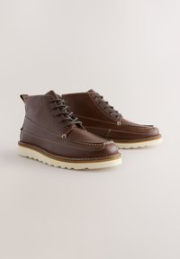 Ruskeat nahkasaappaat, joissa nyöritys, valkoinen ompelu ja kontrastivärinen pohja. Varustettu pyöreällä kärjellä ja nilkan korkeudella.