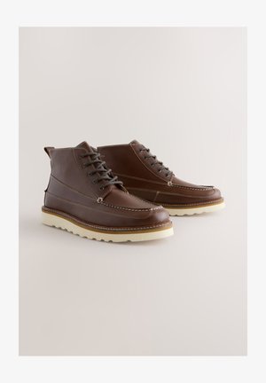 Bruine leren laarzen met vetersluiting, witte stiksels en een contrasterende zool. Heeft een ronde neus en een enkelhoogte.