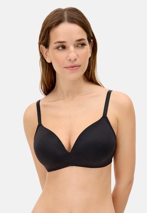 Mujer de piel clara y cabello castaño, viste un sujetador negro sencillo con tirantes finos, mirando ligeramente hacia su izquierda contra un fondo neutro.