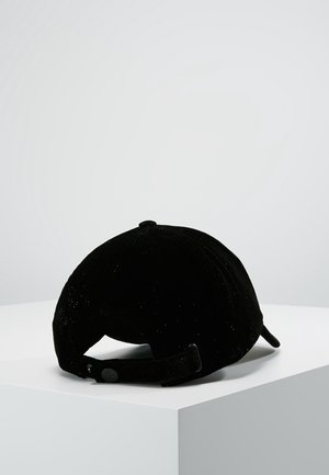 Musta samettinen lippalakki säädettävällä remmillä, valkoisella pinnalla vaalean harmaan yksivärisen taustan edessä.