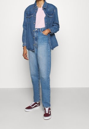 Veste en denim bleu moyen avec deux poches poitrine, portée sur un T-shirt rose clair. Jean bleu délavé à jambes droites et baskets bordeaux.