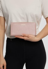 Portafoglio in pelle rosa con una finitura testurizzata, logo dorato impresso sulla parte anteriore, chiusura con zip e forma rettangolare elegante.