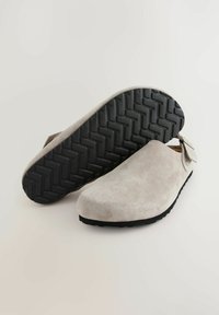 Sabots en daim gris clair avec une forme courbée, une sangle ajustable et une semelle en caoutchouc noir texturé présentant un motif en chevrons pour une meilleure adhérence.