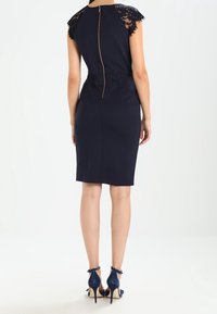Navy sheath dress with lace cap sleeves, featuring a center back zipper. Worn with navy high-heeled shoes. Smooth fabric texture.