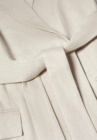 Beżowy płaszcz typu wrap o fakturowanym wyko�ńczeniu, z wiązanym paskiem, bocznymi kieszeniami oraz wyraźnie zarysowanym kołnierzem.