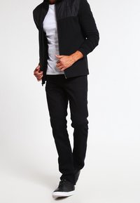 Černá bunda na zip s prošívanými detaily na ramenou, bílé tričko, černé kalhoty a černé tenisky. Jednoduchý, přiléhavý střih.