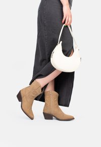 Botas de cowboy de ante marrón con un tacón bajo y diseño bordado. Combinadas con un bolso blanco en forma de media luna. Falda de mezclilla oscura.