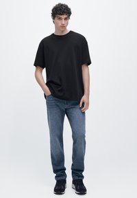 Svart t-shirt med rund hals och korta ärmar, kombinerad med ljusblå denimjeans. Modellen bär svarta sneakers och står mot en enfärgad bakgrund.