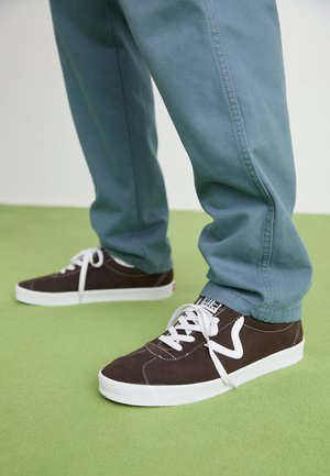 Personne portant des baskets marron foncé avec des lacets blancs et un pantalon bleu debout sur un sol vert.