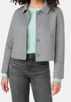 Kvinde iført en grå knappet jakke over en lyse mintgrøn strikket sweater og sorte jeans med små sølvpynt.