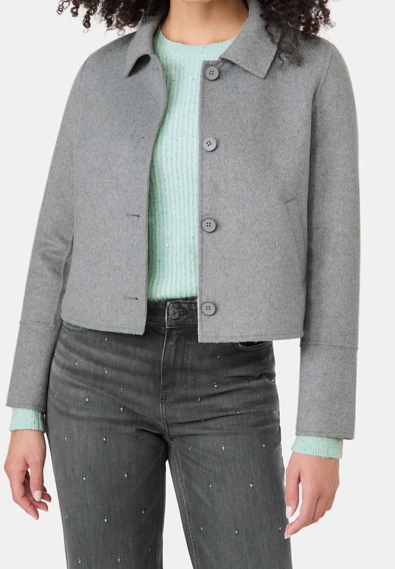 Femme portant une veste grise boutonnée sur un pull en maille vert menthe clair et un jean noir avec de petites décorations argentées.