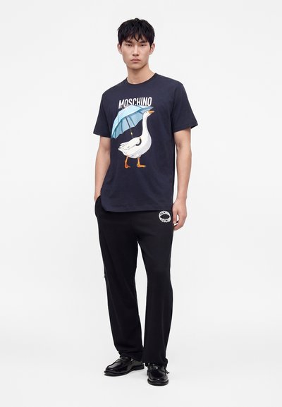 Νέος άνδρας φοράει μπλε σκουρόχρωμο T-shirt Moschino με μια χήνα που κρατάει μια μπλε ομπρέλα και μαύρο παντελόνι με λευκό λογότυπο.