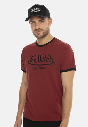 Homme portant une casquette noire Von Dutch et un t-shirt rouge Von Dutch California avec des bordures noires sur les manches et le col.