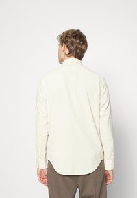 Camicia a maniche lunghe beige chiaro con colletto, tessuto testurizzato, orlo arrotondato e polsini con bottoni. Design minimalista, finitura liscia.