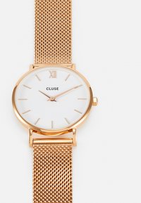 Montre à bracelet en maille rose gold avec un cadran blanc, des marqueurs d'heure minimalistes, et des aiguilles des heures et des minutes en rose gold. La marque "CLUSE" est présente de manière centrale.