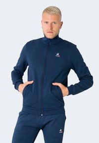 Sudadera de cremallera azul marina con mangas largas, dos bolsillos frontales y puños acanalados. Incluye un pequeño logo en el pecho. Combínala con pantalones a juego.