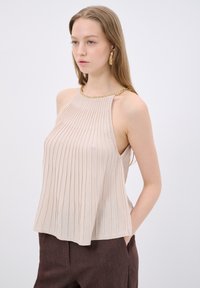 Beige mouwloze top met verticale ribbelstructuur en een gouden kettinghalslijn. Geregeld met donkerbruine broeken, die een losse pasvorm tonen.