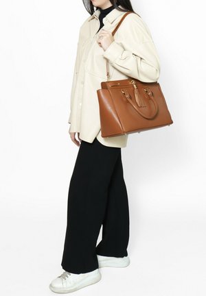 Borsa a tracolla in pelle marrone con design strutturato, manici doppi e dettaglio a nappina. Indossata sopra una camicia crema e pantaloni neri, abbinata a sneaker bianche.