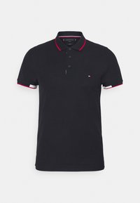 Marinblå polo skjorta med röd och vit randig krage och manschetter, med tre svarta knappar och en liten logotyp på vänster bröst. Tillverkad av bomull.