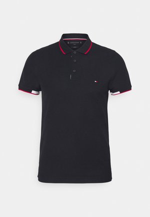 Polo shirt bleu marine avec col et poignets à rayures rouges et blanches, comportant trois boutons noirs et un petit logo sur la poitrine gauche. Matériau en coton.