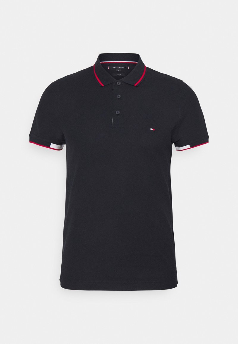 Marinblå polo skjorta med röd och vit randig krage och manschetter, med tre svarta knappar och en liten logotyp på vänster bröst. Tillverkad av bomull.