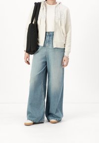 Osoba v světle béžové mikině GAP, bílém tričku, širokých vybledlých modrých džínách, bílých botách a s velkou černou kabelem na rameno.