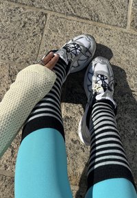 Persona, kas valkā melnas un baltas svītrainas ceļgalu zeķes, baltas sporta kurpes ar zvaigžņu rakstu, zilas legingus un teksturētu krēmkrāsas piedurkni uz akmens bruģa.