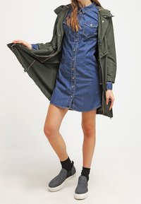 Denim-paita mekko, jossa on nappikiinnitys ja kaulus, kerroksittain vihreän vedenpitävän parkan alla. Harmaat liukuhihnatossut mustilla sukilla.