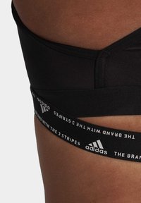 Črna atletska modrček, ki se ponaša z gladkim materialom, debelim črnim pasom z belim besedilom in belim logotipom Adidas. Poudarjen z brezšivno zasnovo.