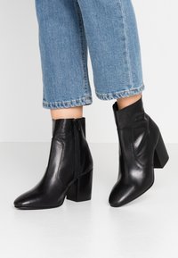 Piernas vestidas con jeans cortos de color azul y botas de cuero negras hasta el tobillo con cremalleras laterales y tacones bloque sobre un suelo blanco.