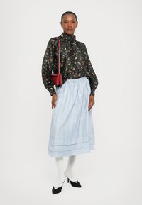 Blusa nera floreale con maniche lunghe, gonna plissettata azzurra chiara, collant bianchi e tacchi neri, abbinati a una piccola borsa rossa.