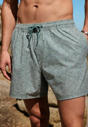 Man wearing teal and white geometric patterned swim shorts with a drawstring, standing outdoors near rocks under clear sky.