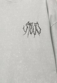 Felpa grigia chiara con un logo ricamato scuro e testurizzato in un carattere frastagliato sull'area sinistra del petto. Tessuto morbido e leggermente usurato.