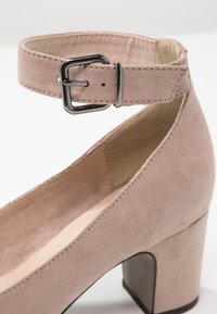 Chaussure à talon haut en daim beige avec bride de cheville et boucle argentée, bout arrondi et talon bloc épais.