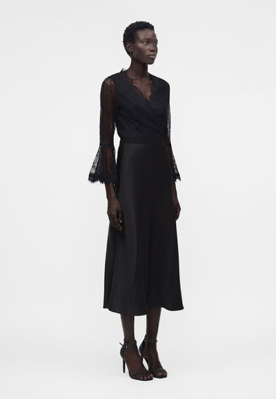 Γυναίκα στέκεται φορώντας μαύρο μακρυμάνικο τοπ από δαντέλα με φαρδιά μανίκια, μαύρη μίντι φούστα και μαύρα ψηλοτάκουνα σανδάλια μπροστά σε απλό λευκό φόντο.