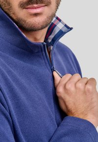 Sudadera navy con cremallera y un tejido texturizado, con un cuello interior a cuadros. Las características incluyen un cuello alto y una cremallera visible en la mitad.