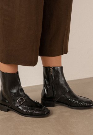 Botines de cuero negro brillante con cremallera lateral y detalle de hebilla, usados con pantalones cortos marrones sobre un suelo beige.