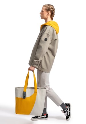 Gray oversized jacket with yellow hood, light gray pants, black shoes. Carrying a large tote bag with gray and bright yellow sections.