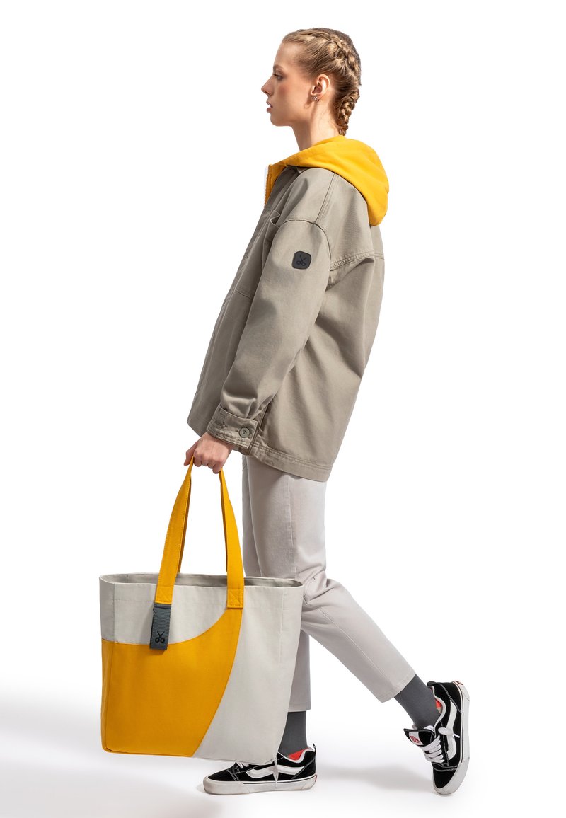 Grijze oversized jas met gele capuchon, lichtgrijze broek, zwarte schoenen. Draagt een grote tas met grijze en felgele secties.