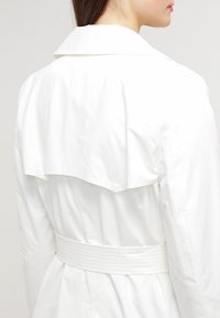 Vit trenchcoat med en utsvängd bakkrage, bälte i midjan och slät textur. Har en enkel, strukturerad design.