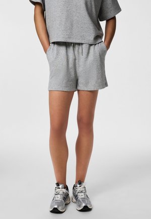 Persoon draagt grijze casual korte broek en bijpassend losvallend kortärmelig shirt, met handen in de zakken en zilveren sneakers, tegen een effen achtergrond.