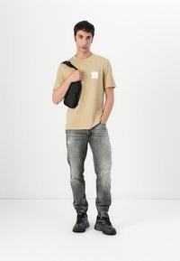 T-shirt en coton beige avec un petit patch blanc, associé à un jean en denim gris avec des détails déchirés. Sac à bandoulière noir et baskets.