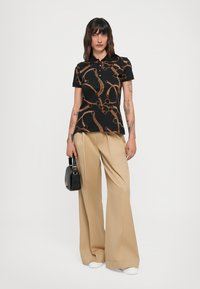 Maglietta polo nera con motivo a catena marrone, abbinata a pantaloni beige a gamba larga. Accessoriata con una piccola borsa nera.