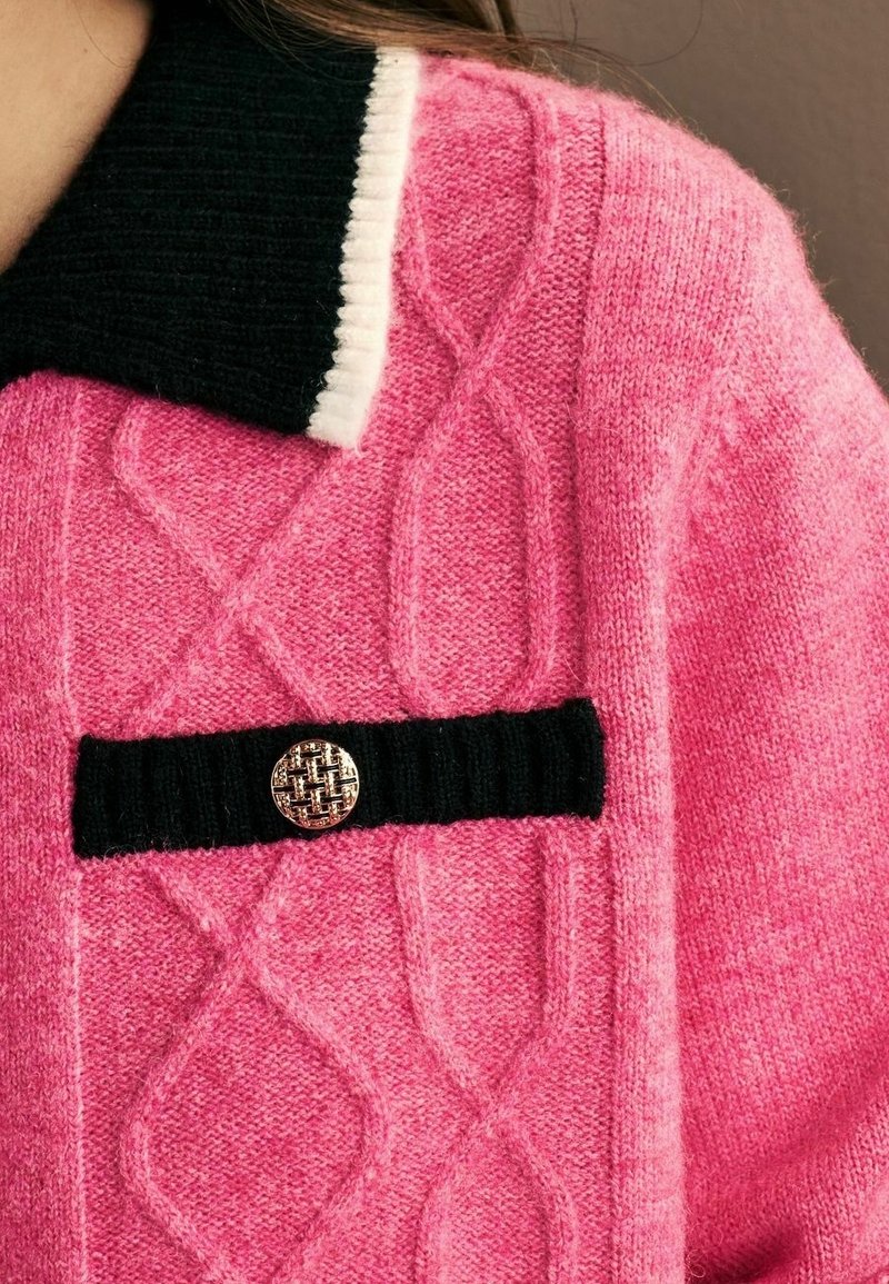 Pull en maille torsadée rose avec détail de col noir et blanc et un patch côtelé noir avec un bouton texturé doré.