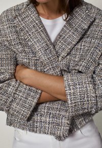 Personne portant une veste en tweed texturée et multicolore avec les manches retroussées sur une chemise blanche, bras croisés à la taille.